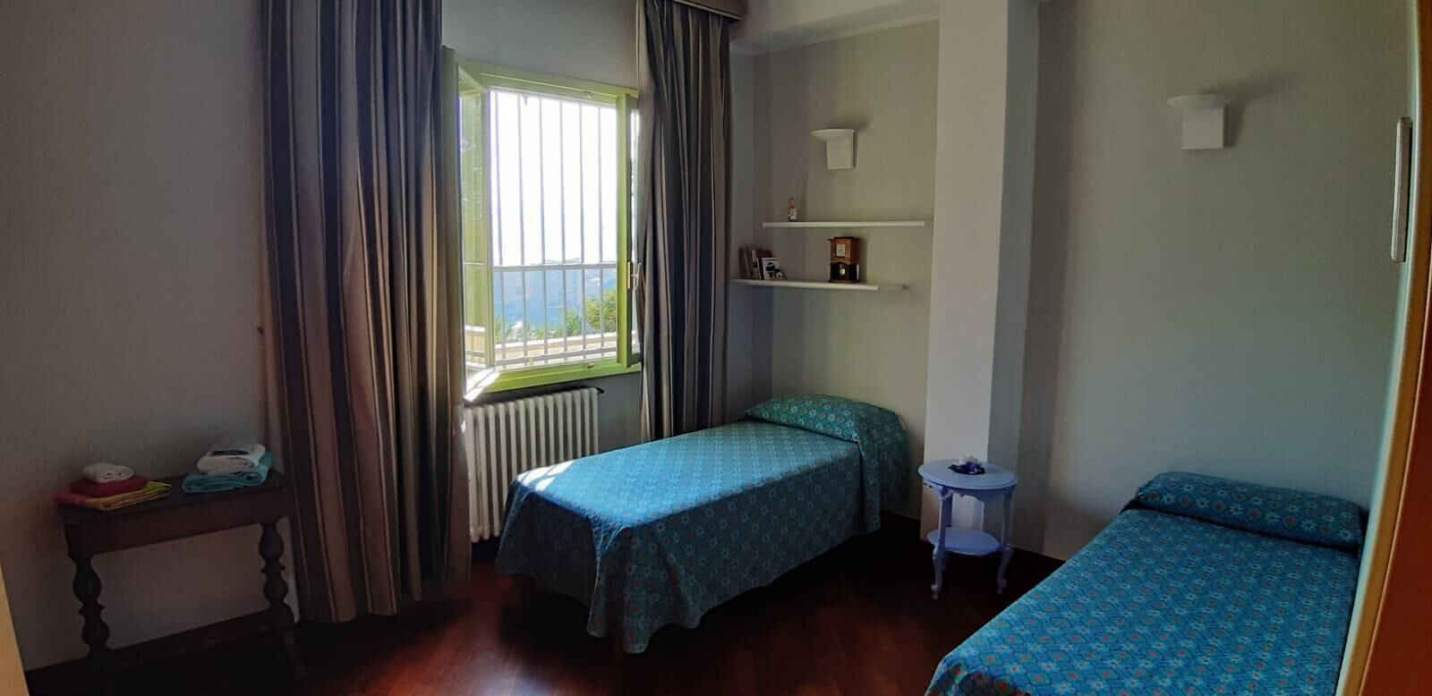 Une petite pièce bien éclairée, avec deux lits simples, une petite table, un radiateur et une fenêtre avec des rideaux.