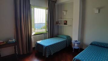 Une petite pièce bien éclairée, avec deux lits simples, une petite table, un radiateur et une fenêtre avec des rideaux.