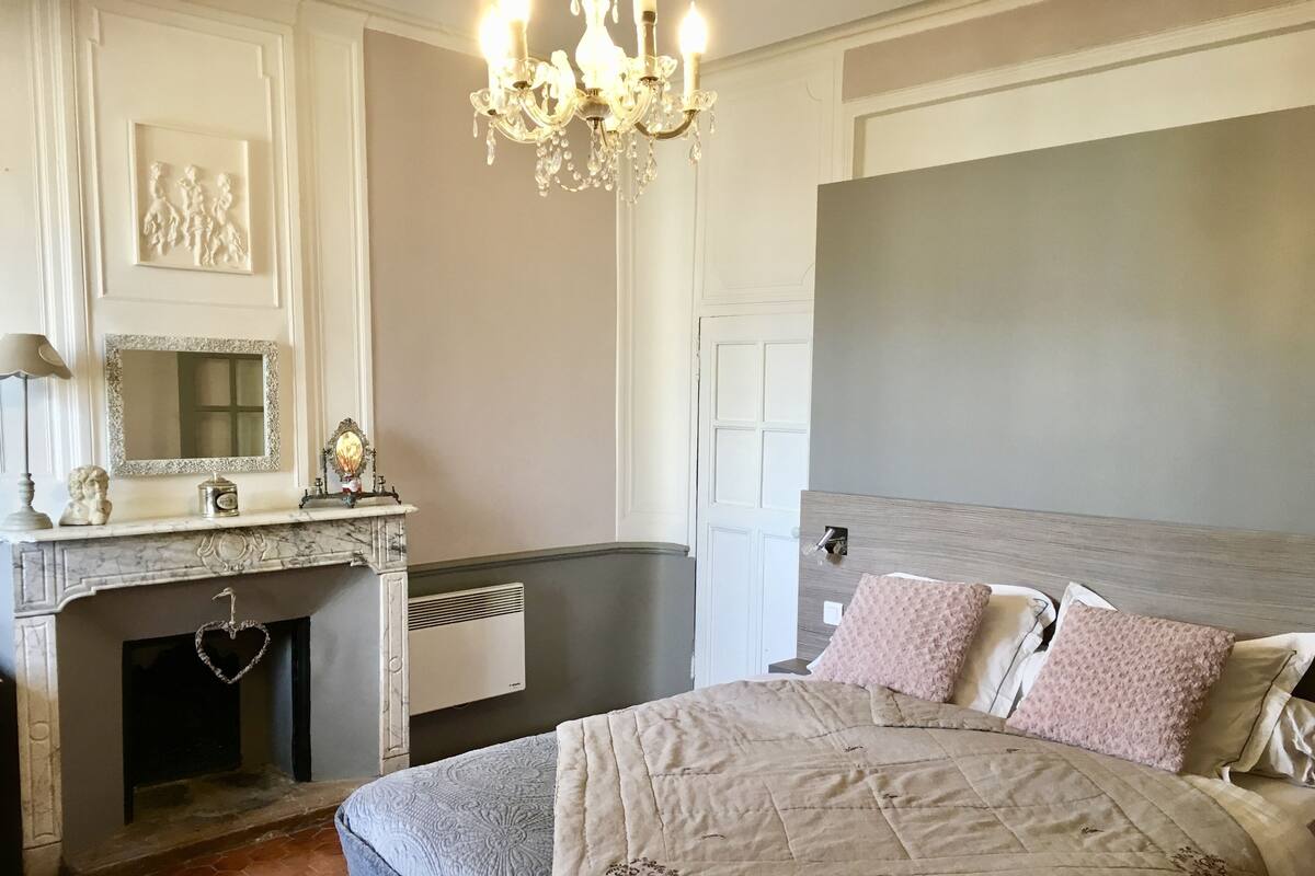 A bedroom with a large bed, a fireplace, a chandelier, and a mirror.