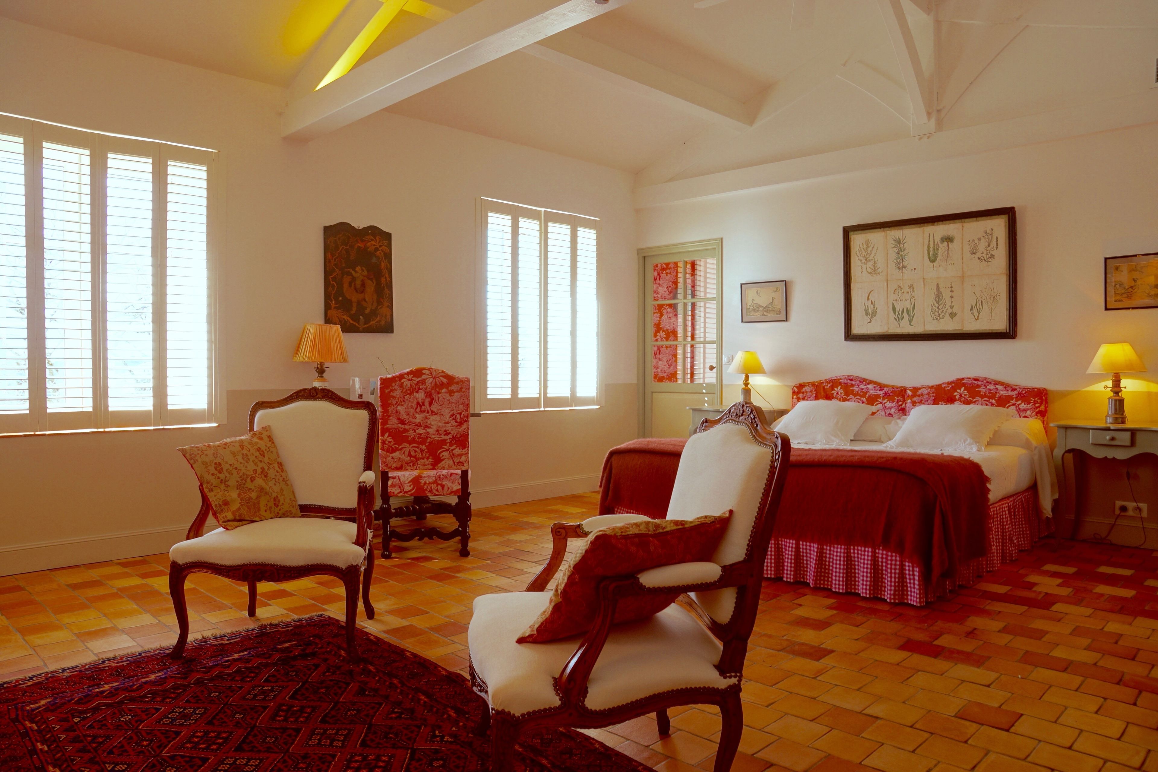 Une pièce spacieuse avec un sol à carreaux, deux lits avec une literie rouge, une chaise en bois, un fauteuil à motifs rouges et blancs, et une grande estampe représentant des plantes accrochée au mur.