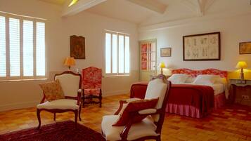 A spacious room with a checkered floor, two beds with red bedding, a wooden chair, a red and white patterned armchair, and a large botanical print on the wall.