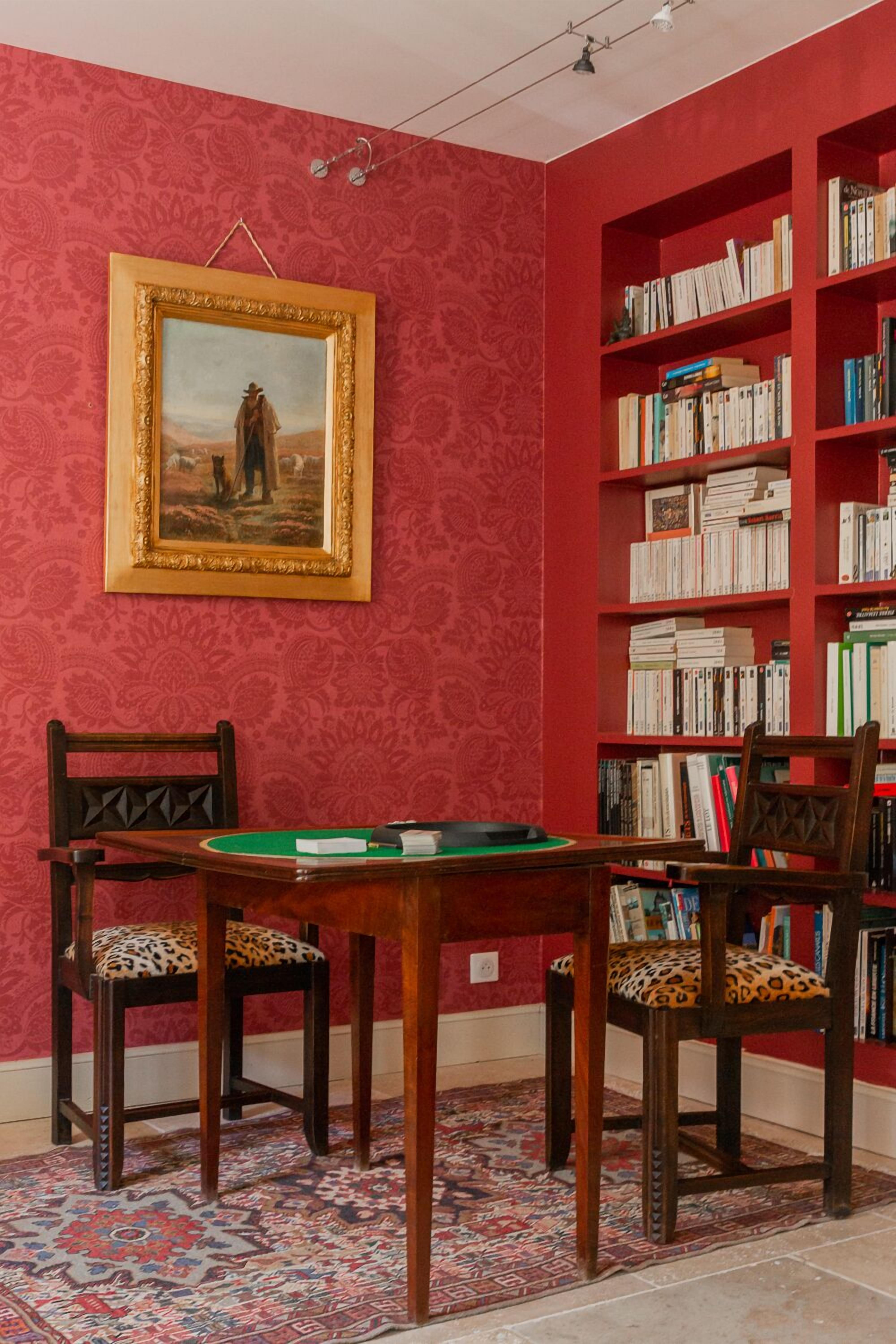 A room with a red patterned wall, a framed painting, a bookshelf, a table with chairs, and a patterned rug.