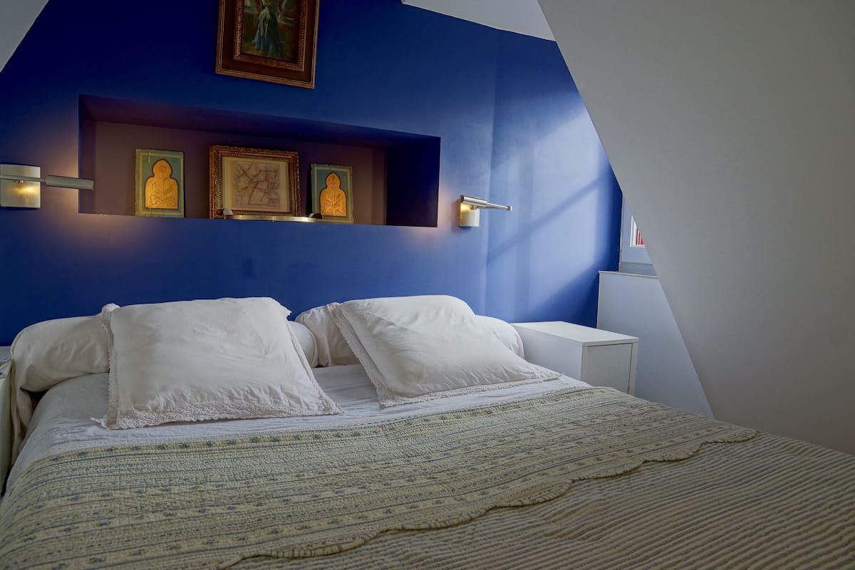 Un dormitorio con cama, almohadas blancas, manta estampada, pared azul decorativa y cuadros enmarcados.