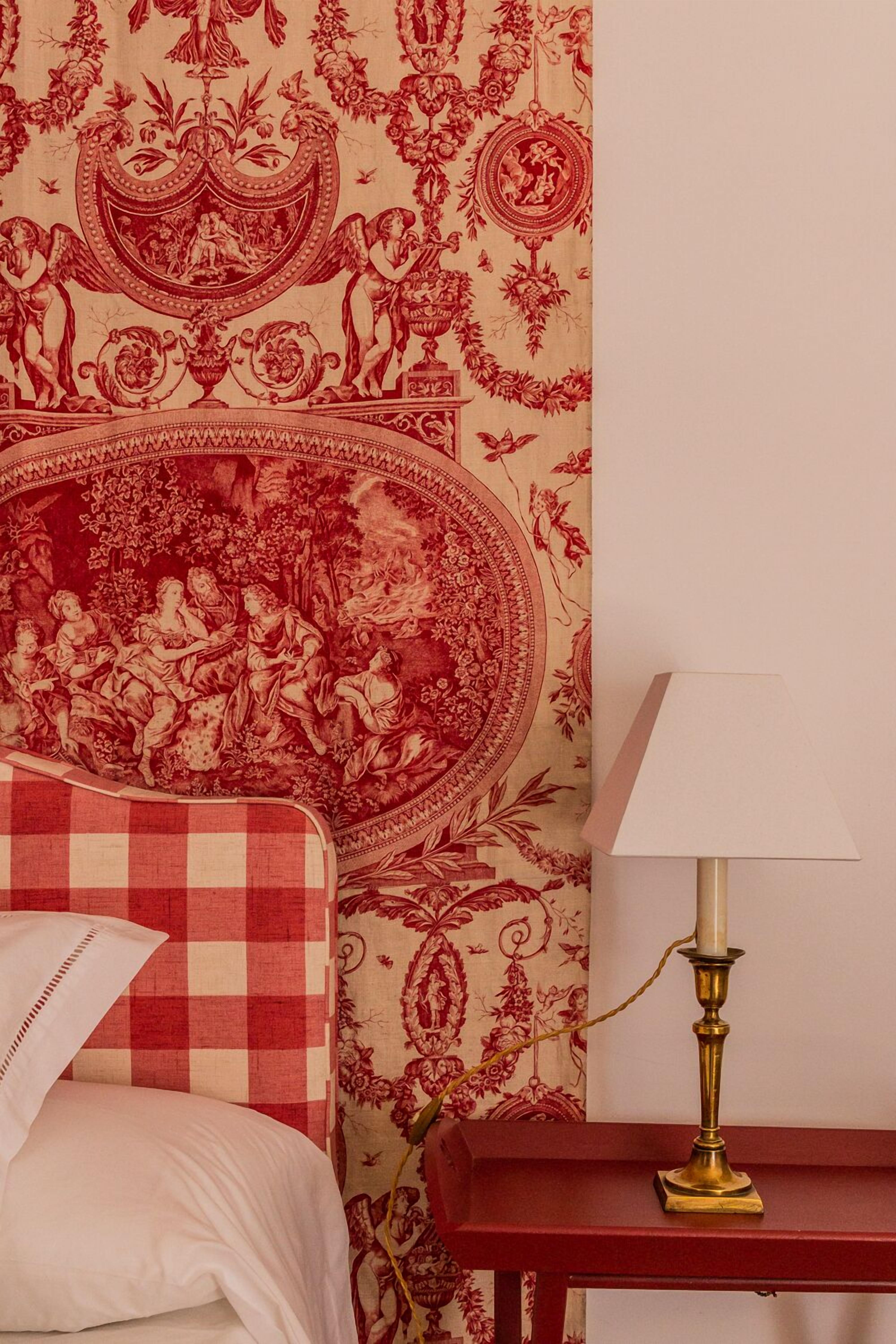 Une chambre à coucher avec une couvre-lit à carreaux, un papier peint rouge et beige à motifs, et une table de chevet avec une lampe.