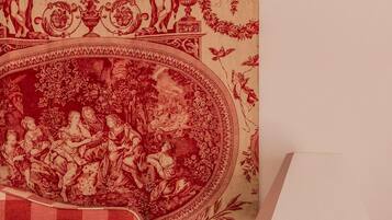 Une chambre à coucher avec une couvre-lit à carreaux, un papier peint rouge et beige à motifs, et une table de chevet avec une lampe.