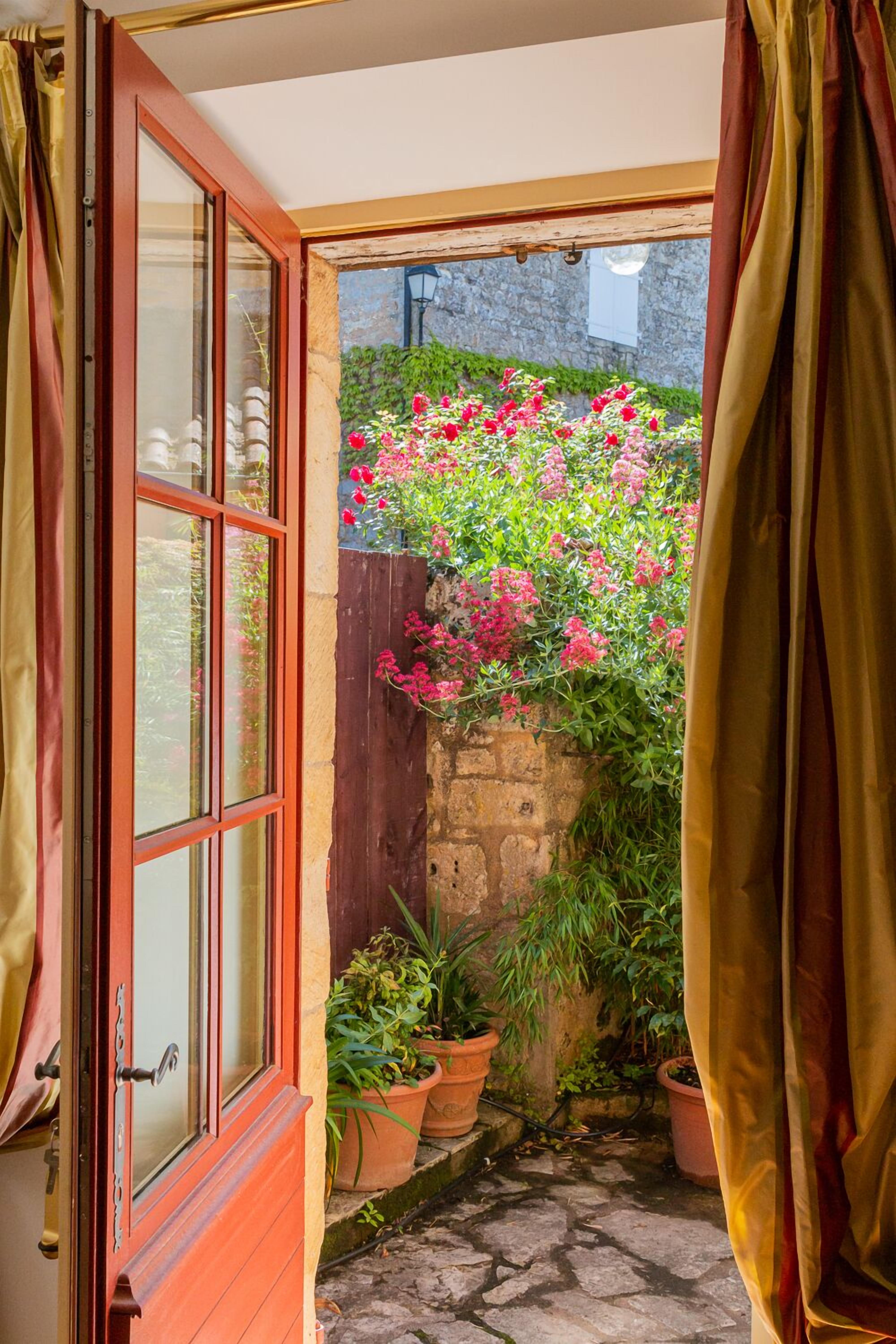 An open wooden door leading to a garden with potted plants and flowering plants.