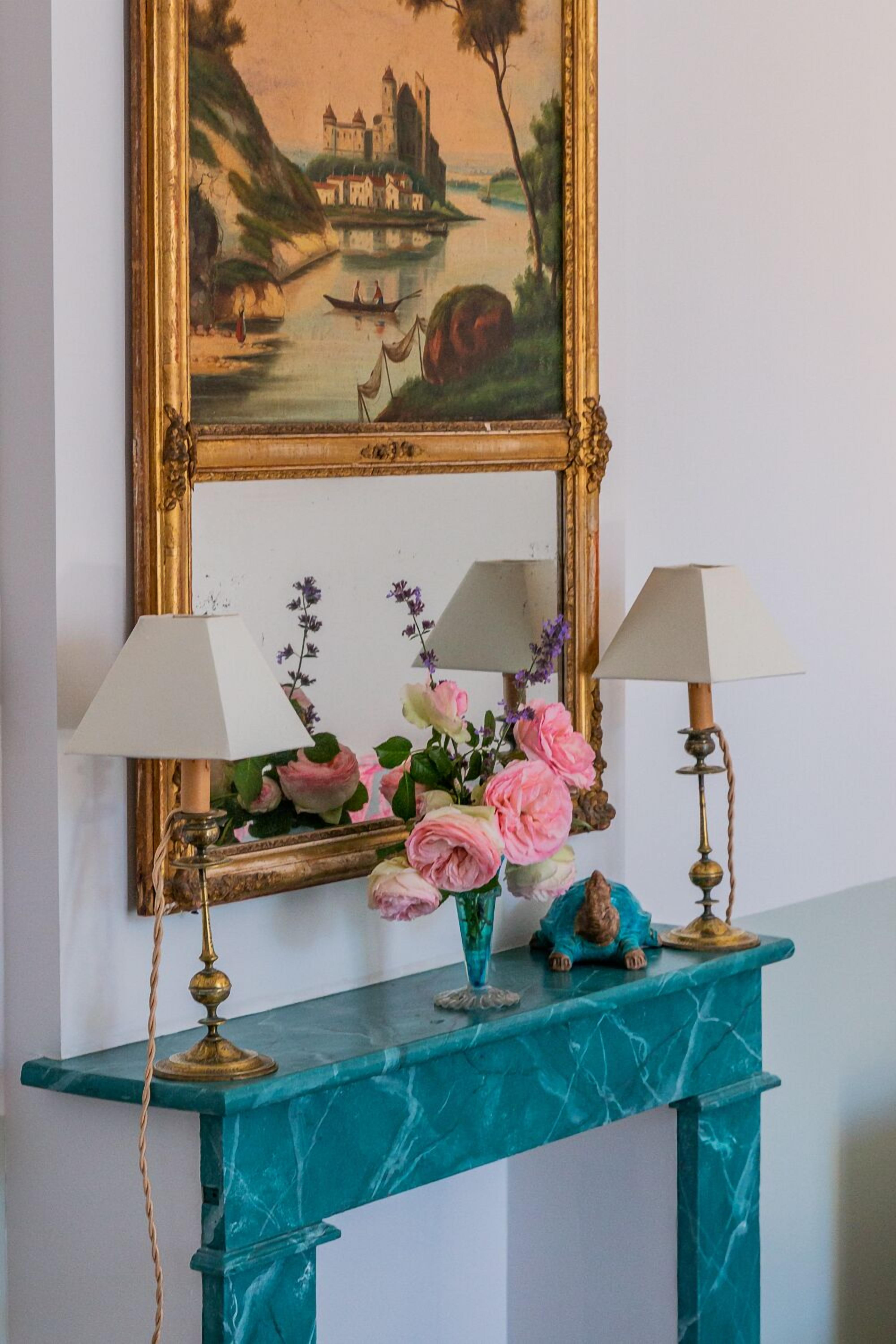 A teal marble console table with two lamps, a vase of pink flowers, and a small figurine.