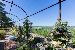 Un jardín con una mesa y sillas, rodeado de plantas en flor y vistas a un valle.