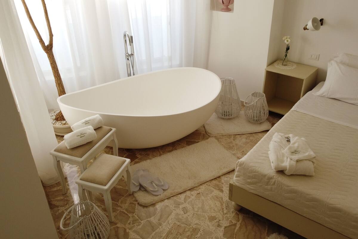 A modern bathroom with a freestanding bathtub, a small bedside table with a vase, and a fluffy rug.