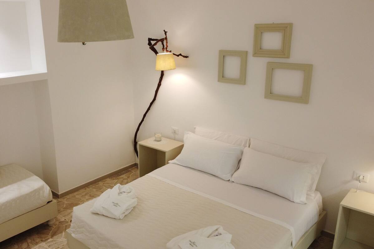 A hotel room with a bed, a bedside table, a lamp, and a window with a plant.