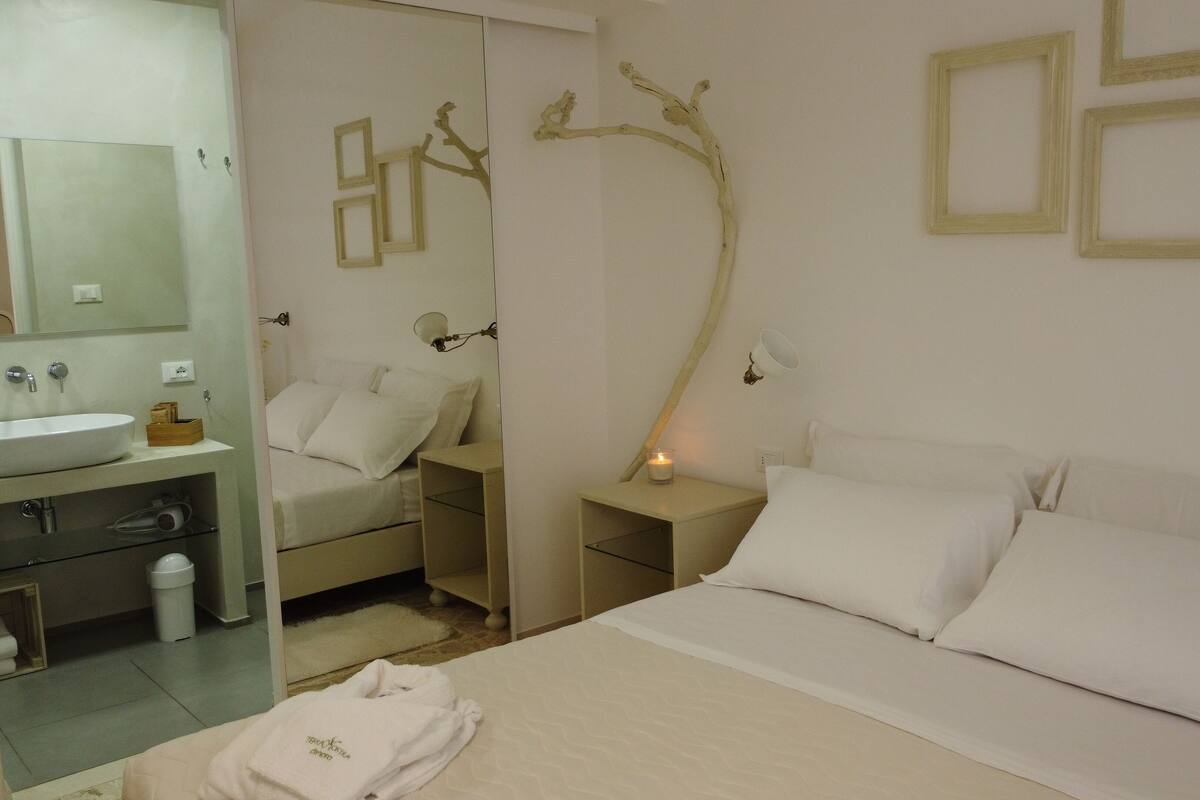 A bedroom with a bed, nightstands, a mirror, and a bathroom with a sink and towel rack.