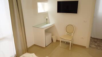 Una habitación pequeña y limpia con un tocador blanco, un espejo, un televisor montado en la pared, una silla blanca y un cesto de basura pequeño.