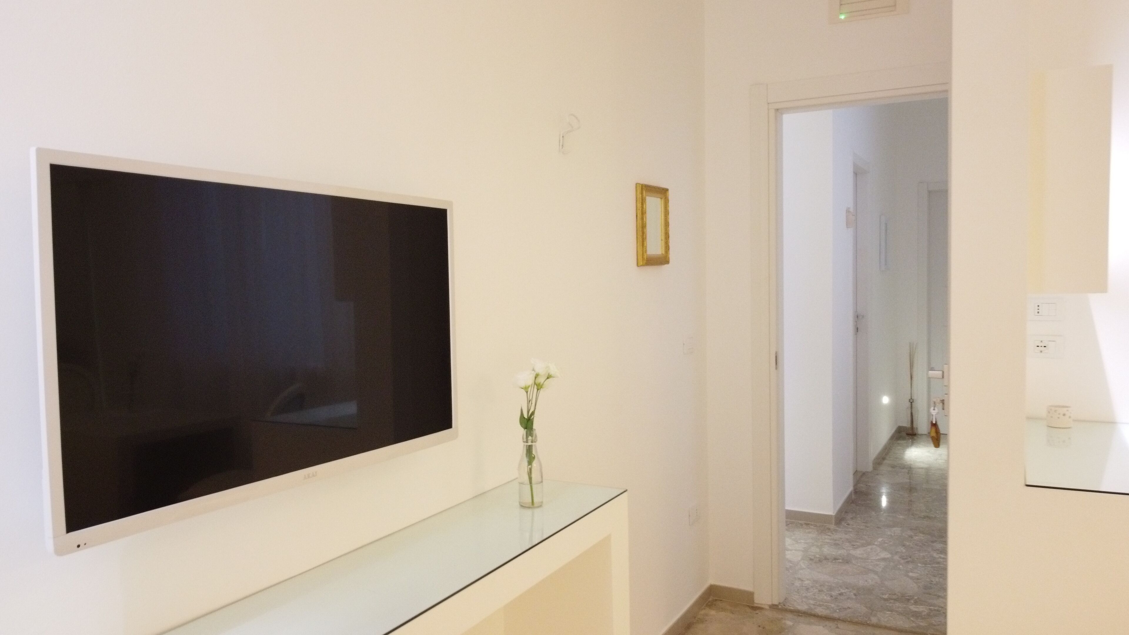 Una habitación moderna con un televisor de pantalla plana grande montado en la pared, una mesa consola de vidrio con un jarrón de flores, y un pasillo que conduce a otra habitación.