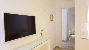 Una habitación moderna con un televisor de pantalla plana grande montado en la pared, una mesa consola de vidrio con un jarrón de flores, y un pasillo que conduce a otra habitación.