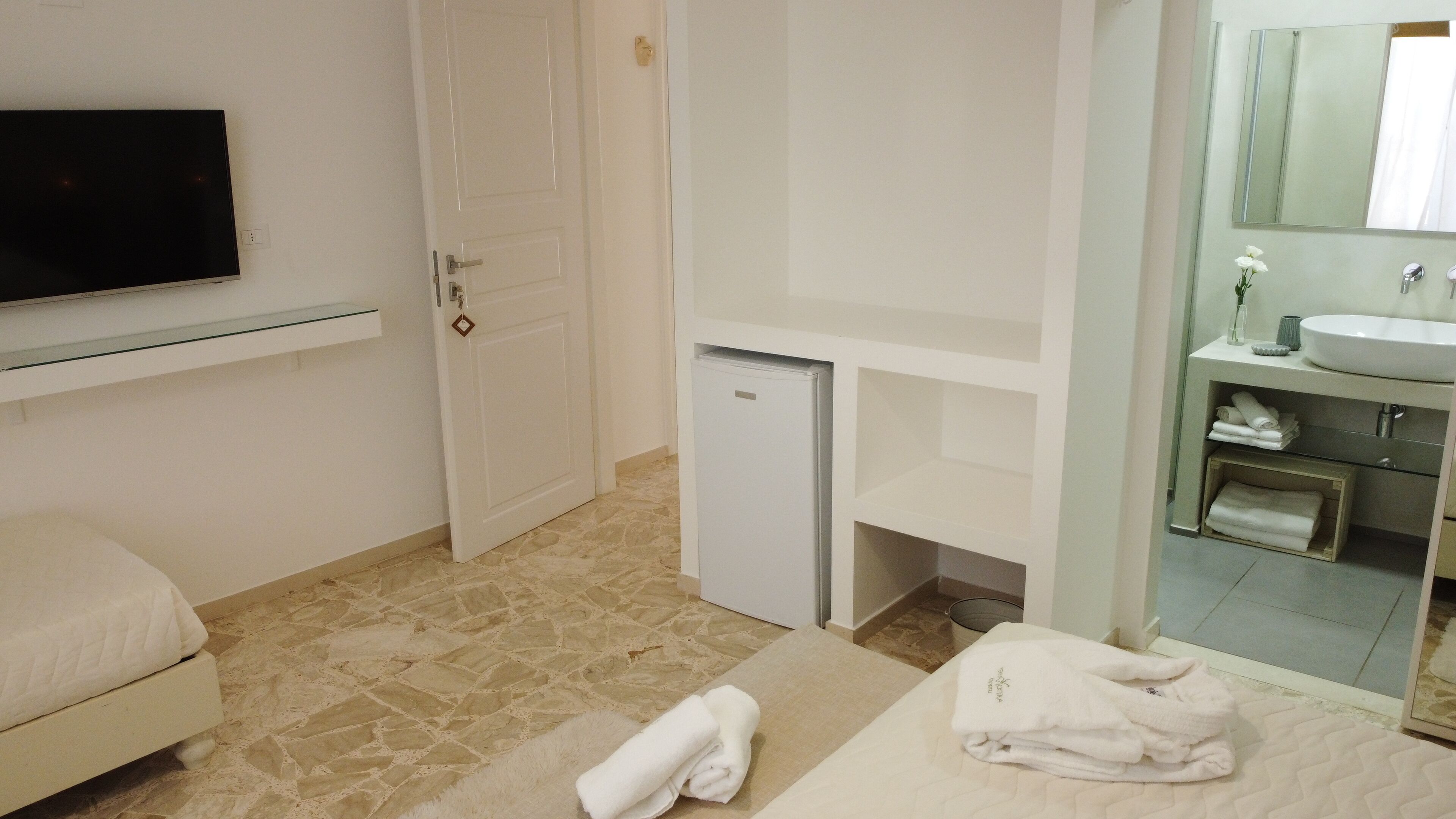 Habitación de hotel con cama, un refrigerador pequeño, baño con lavamanos y espejo, y televisor montado en la pared.