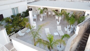 Una terraza en la azotea con sillones blancos, plantas en macetas y un área de estar con mesas y sillas.