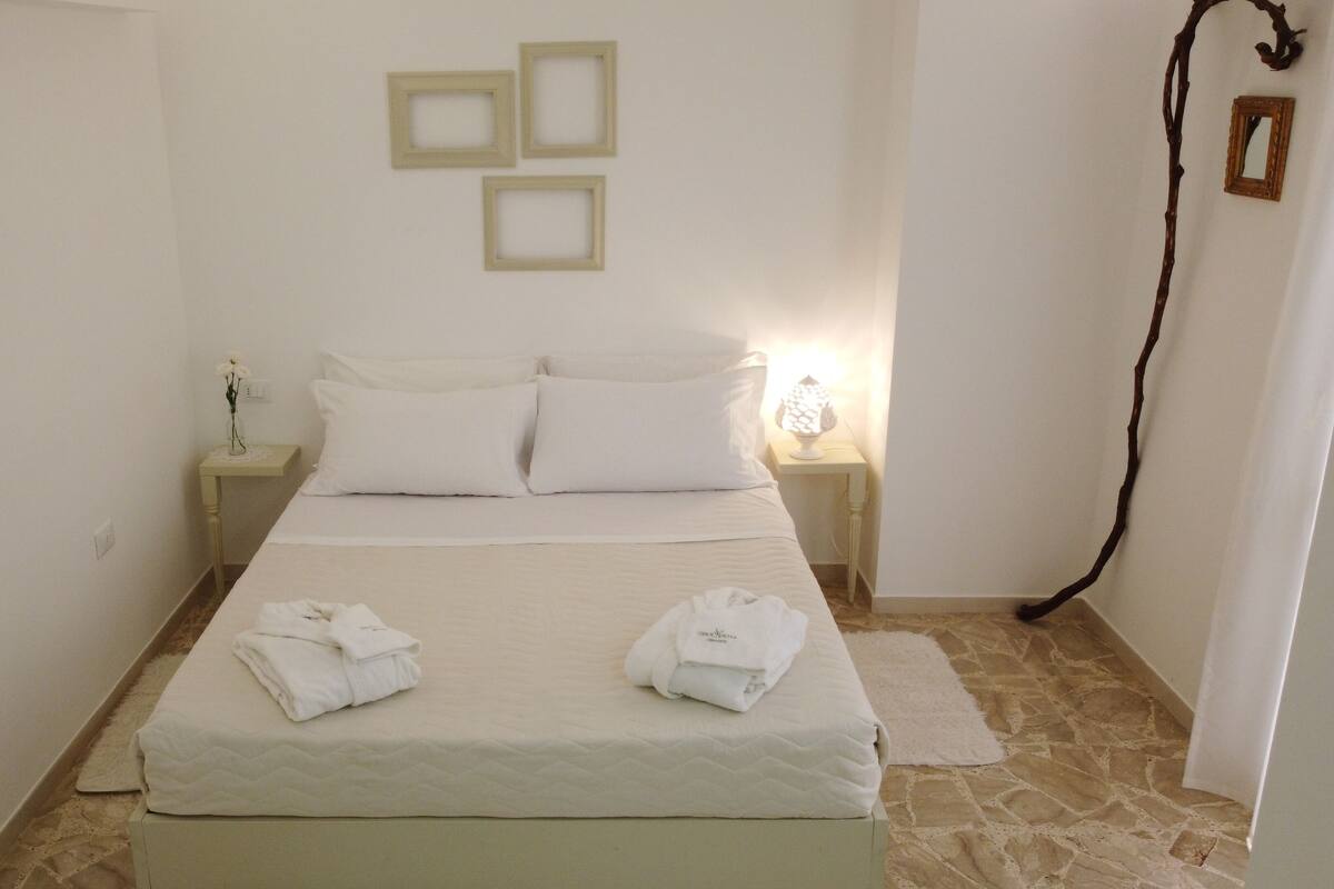 A neatly made bed with two pillows and two folded towels on a white mattress. There are two bedside tables with lamps, a small wall decoration, and a mirror. The room has a stone floor and plain white walls.