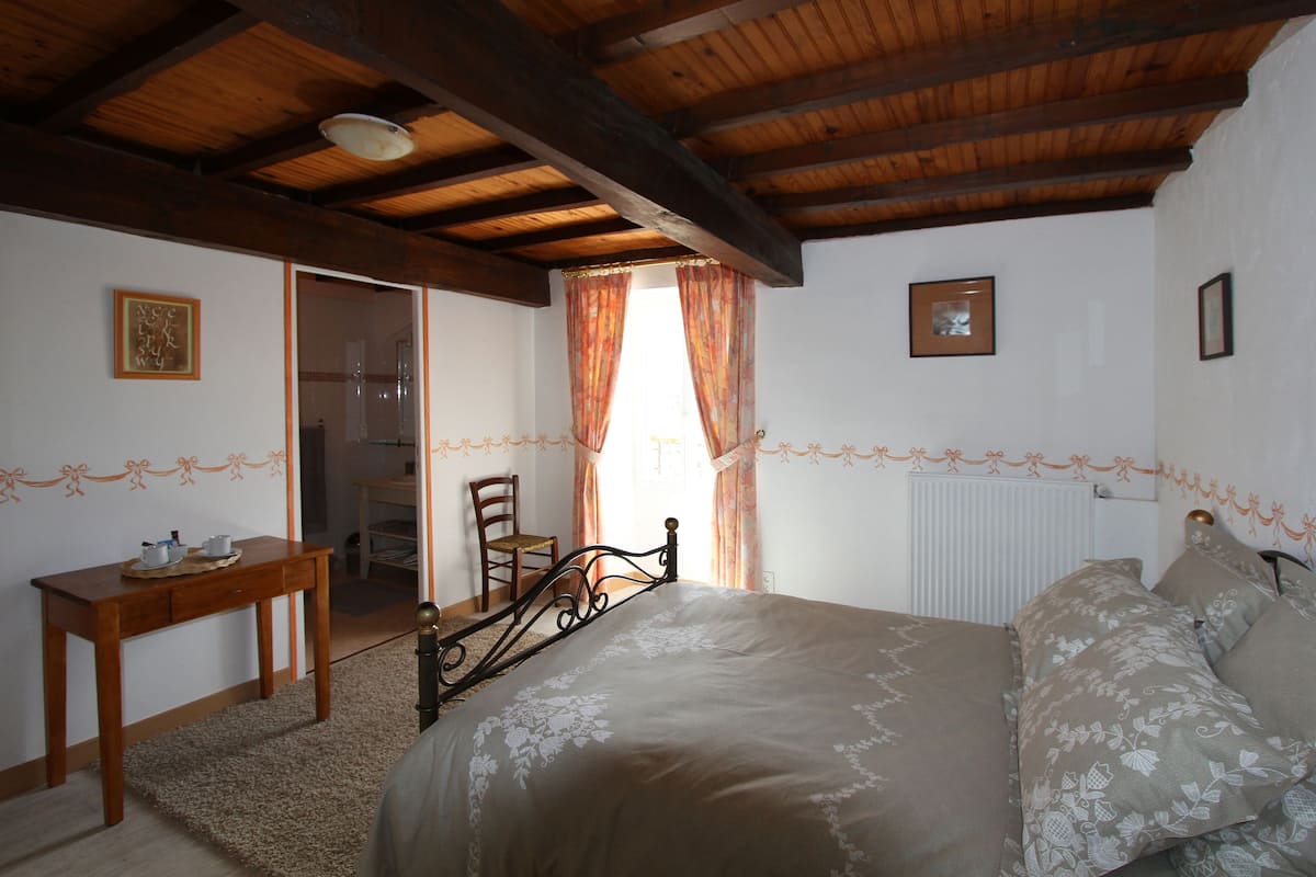 A bedroom with a wooden ceiling, a bed with floral bedding, a wooden nightstand, a chair, and a window with curtains.