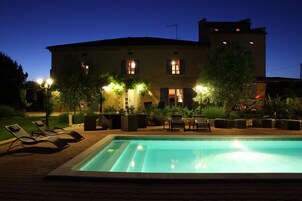 Una piscina con sillas de descanso, un edificio con varias ventanas e iluminación exterior.
