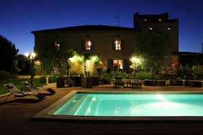 Una piscina con sillas de descanso, un edificio con varias ventanas e iluminación exterior.