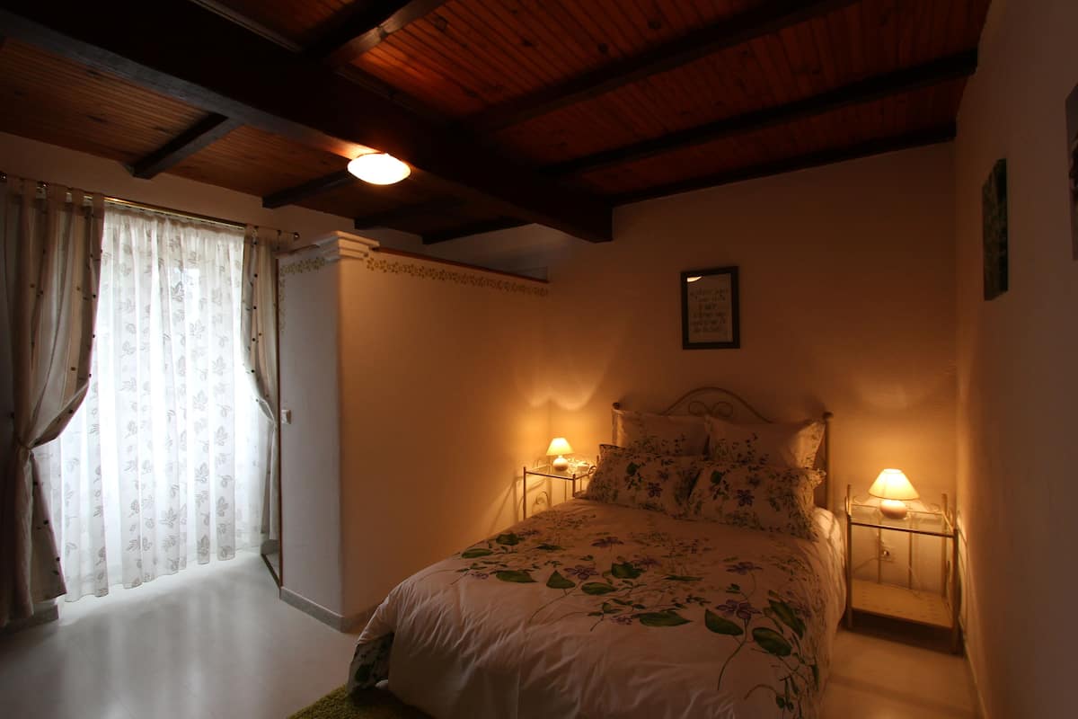 A bedroom with a bed, two bedside lamps, a wooden ceiling, and a window with curtains.