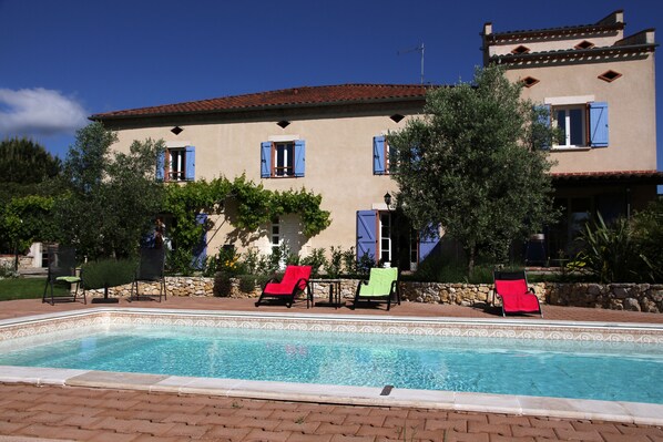 Una piscina con sillones de descanso, un muro de piedra y un edificio con contraventanas azules.