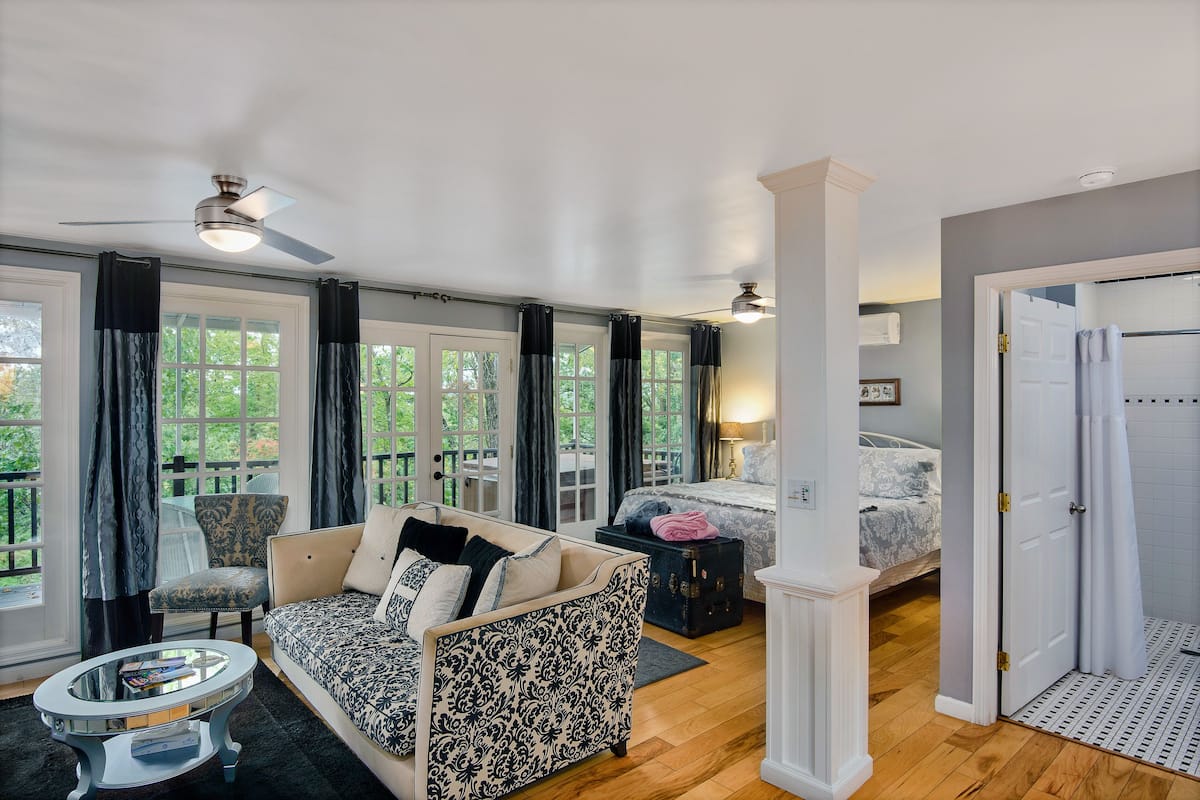 A spacious room with a large bed, a sofa, and a dining table. There are large windows with curtains, a ceiling fan, and a door leading to another room.