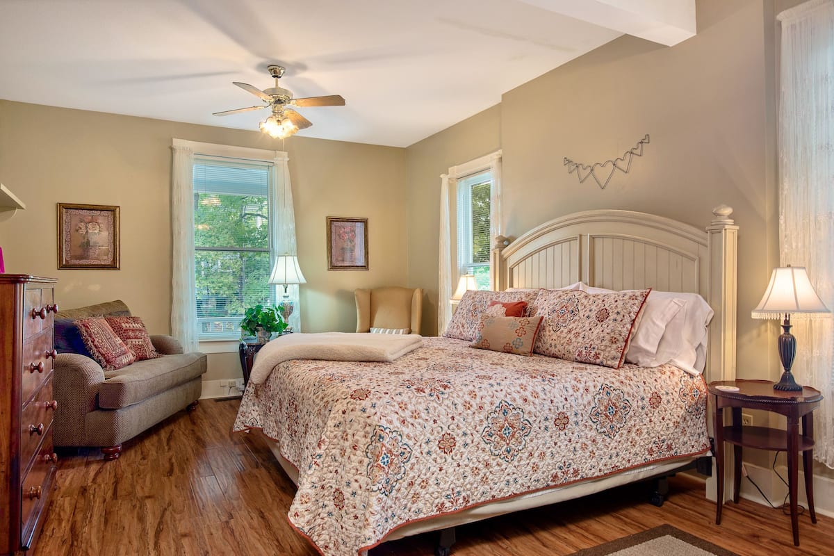 A bedroom with a large bed, a ceiling fan, a dresser, and a window with curtains.