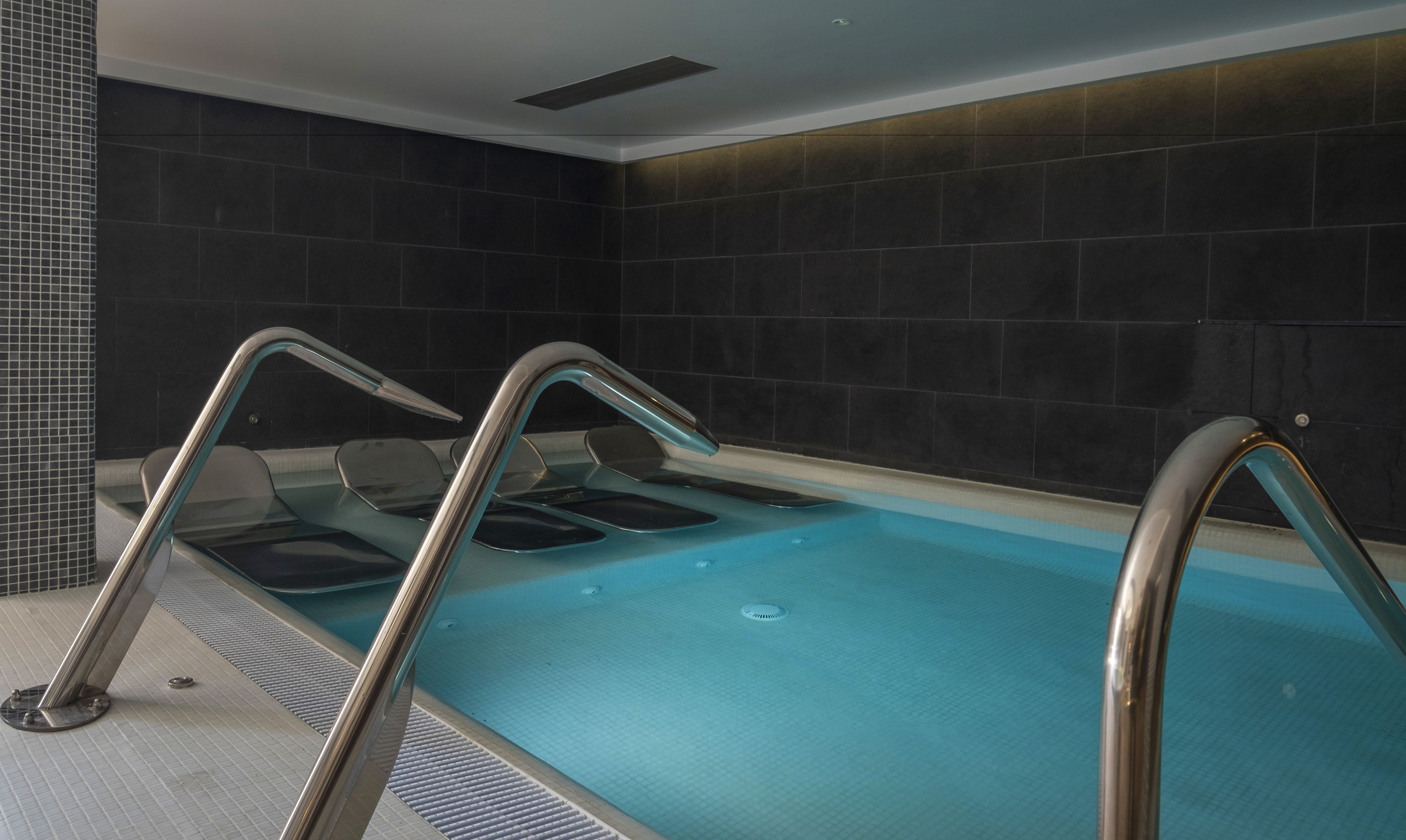 A modern indoor swimming pool with clear water, metal railings, and dark tiled walls.