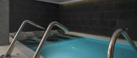 A modern indoor swimming pool with clear water, metal railings, and dark tiled walls.