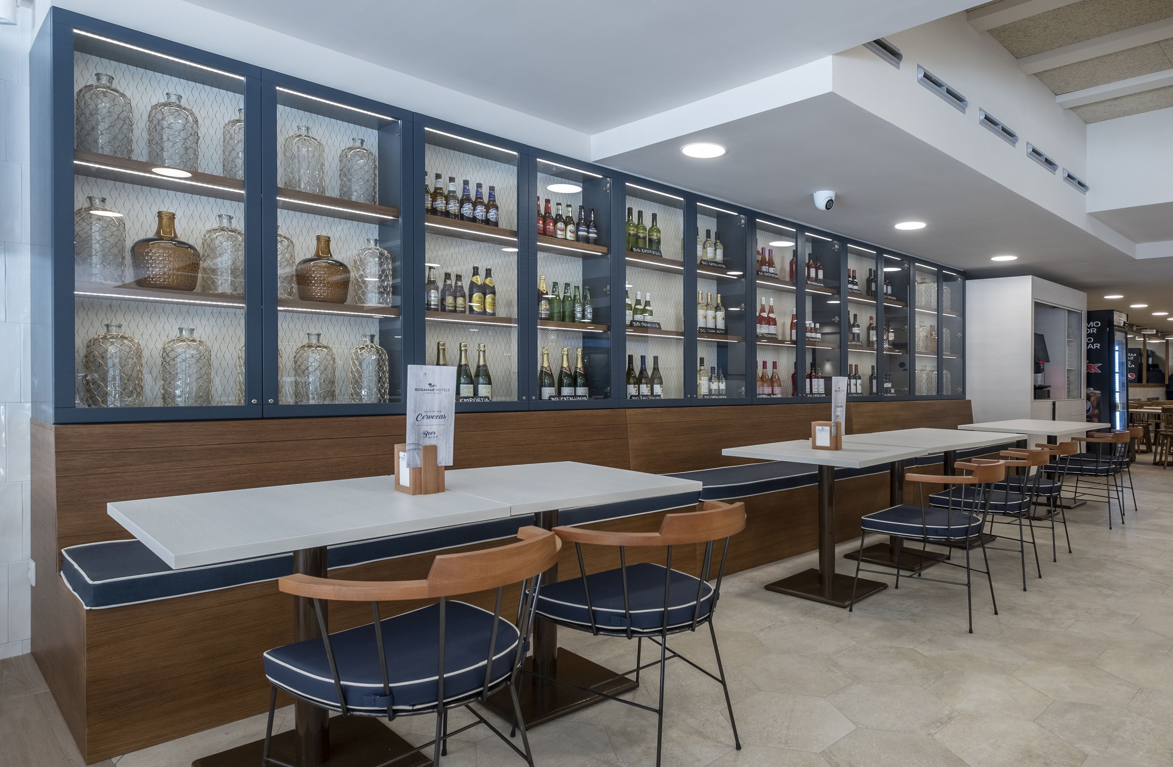 Un restaurante con mesas largas blancas y sillas con reposabrazos azules, una barra de madera y estantes con varias botellas.