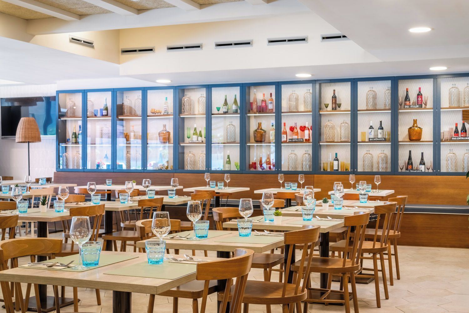 Un restaurante con varias mesas y sillas, una pared de vitrinas de vidrio con botellas y un techo con iluminación empotrada.