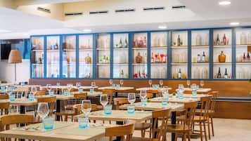 Un restaurante con varias mesas y sillas, una pared de vitrinas de vidrio con botellas y un techo con iluminación empotrada.