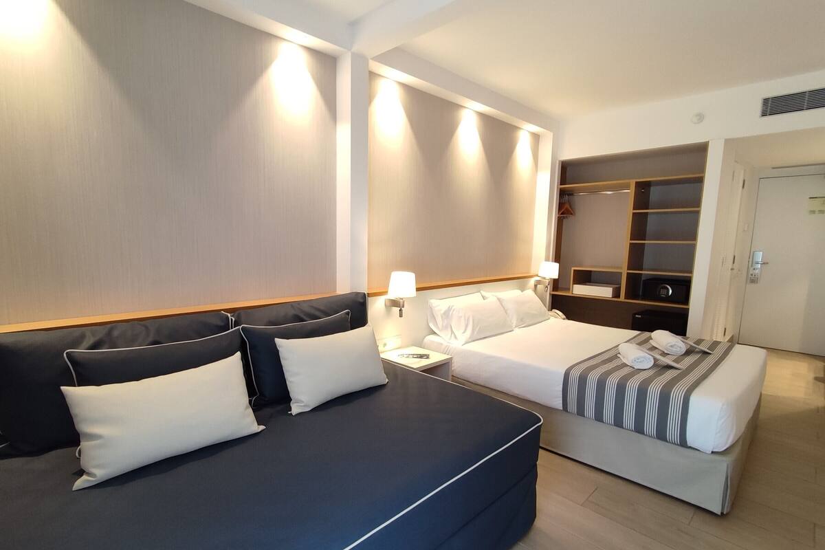 Quarto de hotel com uma cama grande, uma cama menor, mesa de cabeceira, abajur, guarda-roupa e espelho.