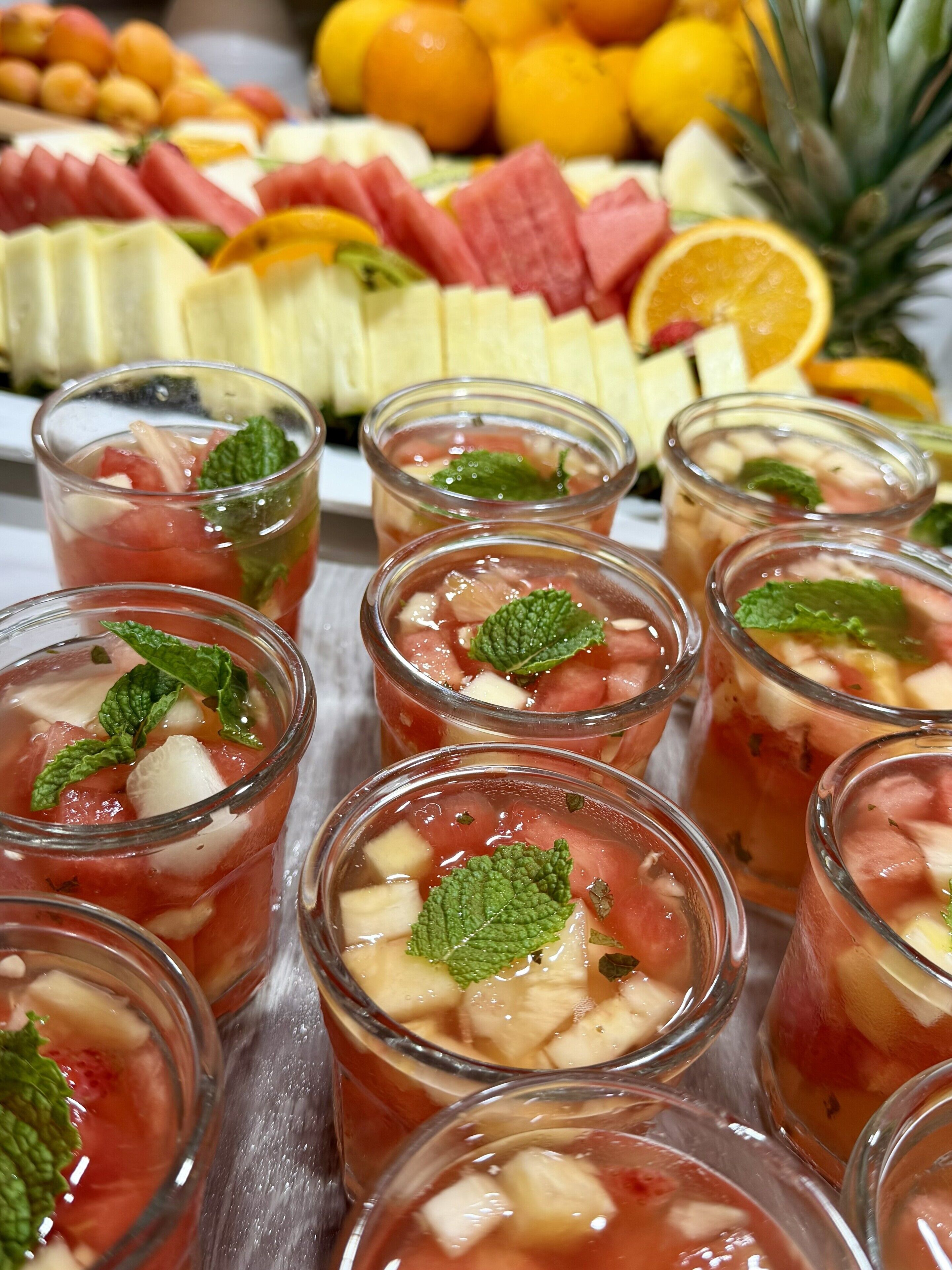 A selection of fruit-based beverages in clear glasses, garnished with mint leaves, with a variety of fruits in the background.