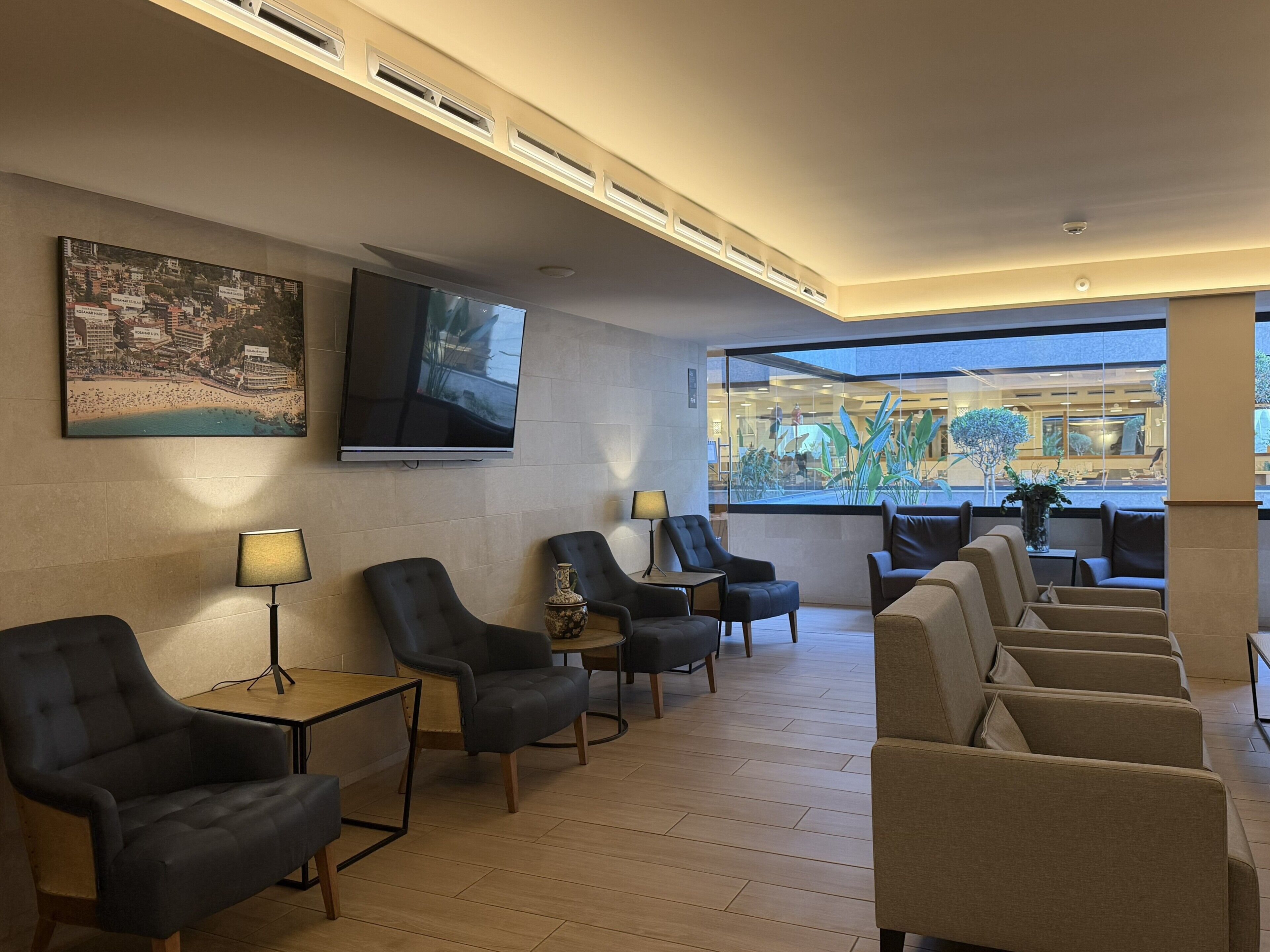 Eine Hotellobby mit einem Flachbildfernseher, einem großen Stadtbildgemälde und einer Sitzecke mit mehreren Stühlen und Tischen.