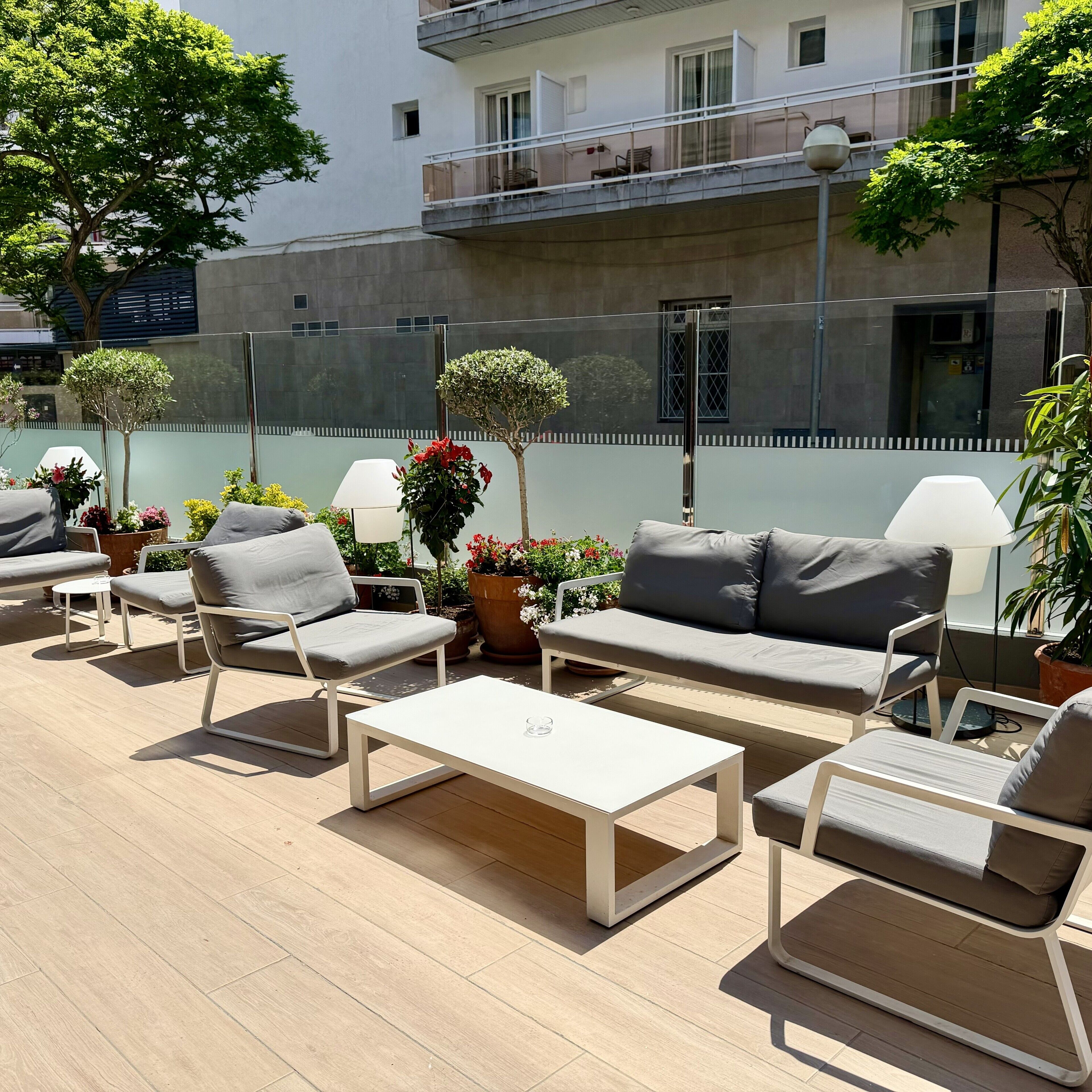 An outdoor seating area with a sofa, chairs, and a coffee table, surrounded by potted plants and a building in the background.