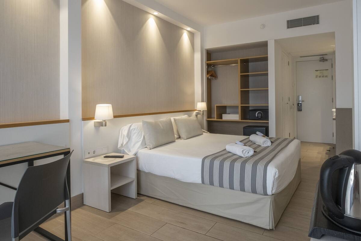 Quarto de hotel com cama, escrivaninha, cadeira, cafeteira e guarda-roupa.