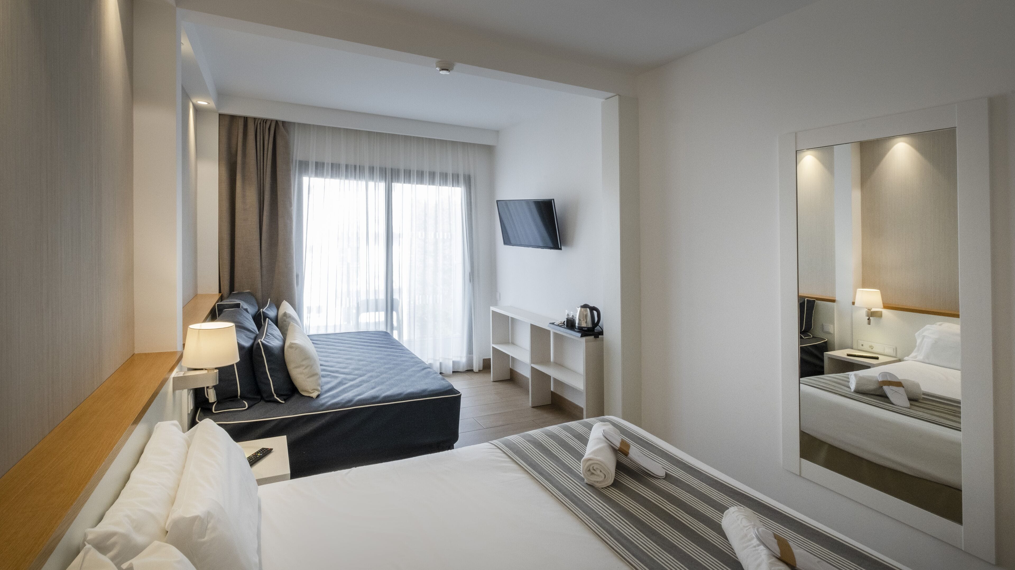 Hotelkamer met een bed, een bureau met lamp, een televisie, een nachtkastje met lamp, een spiegel en een raam met gordijnen.