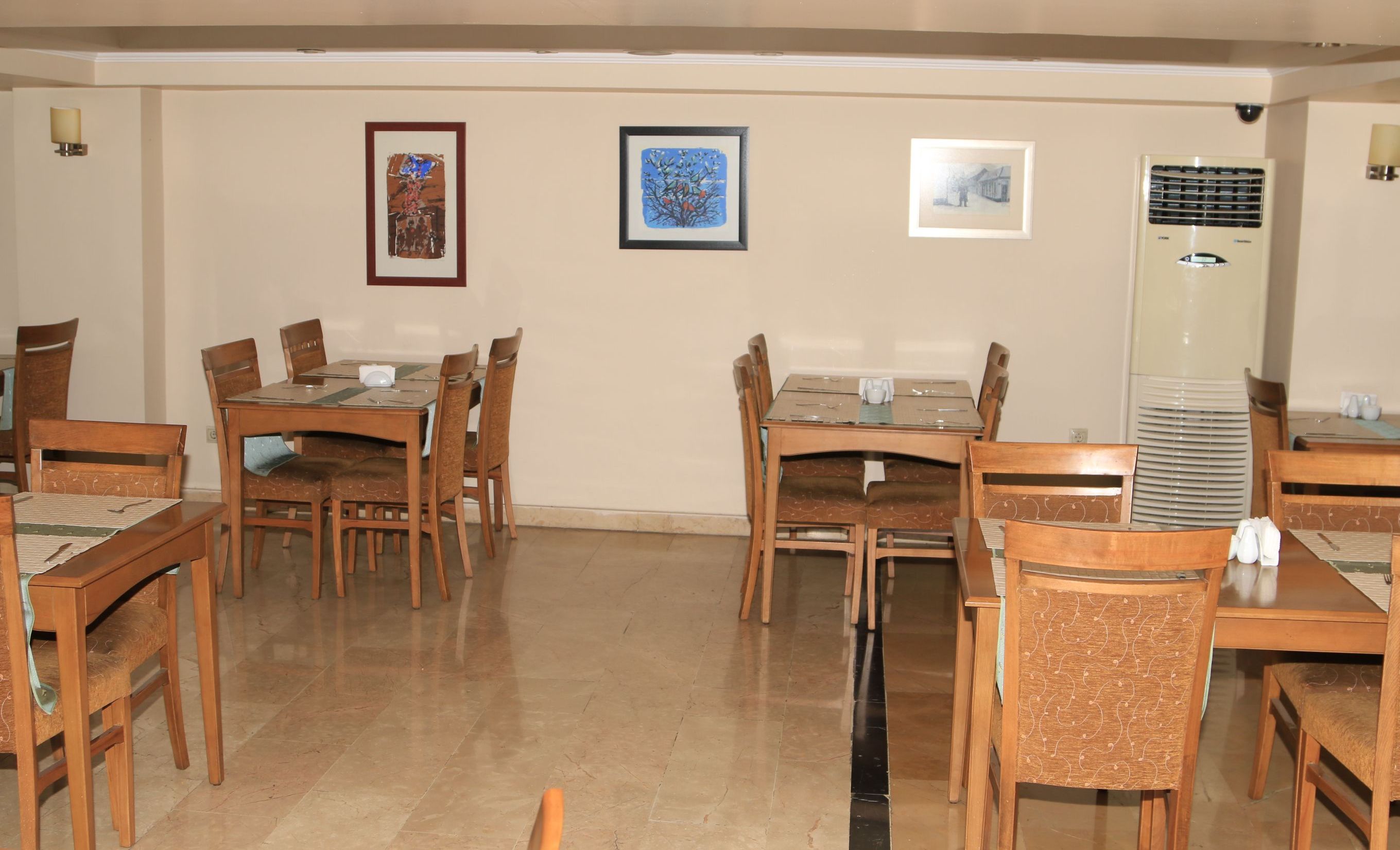 A dining area with wooden tables and chairs, framed artwork on the walls, and a standing air conditioner.