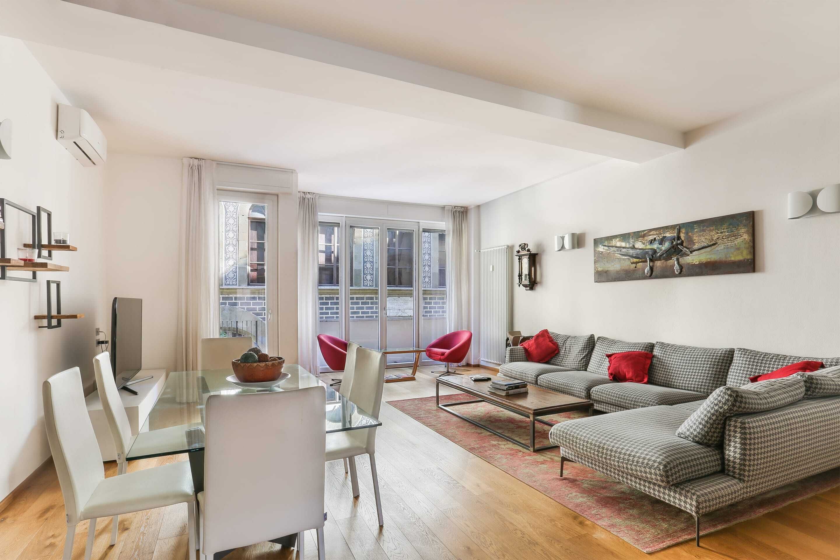Una sala de estar moderna con una mesa de comedor de vidrio, sillas rojas, un sofá gris con cojines rojos y un cuadro en la pared.