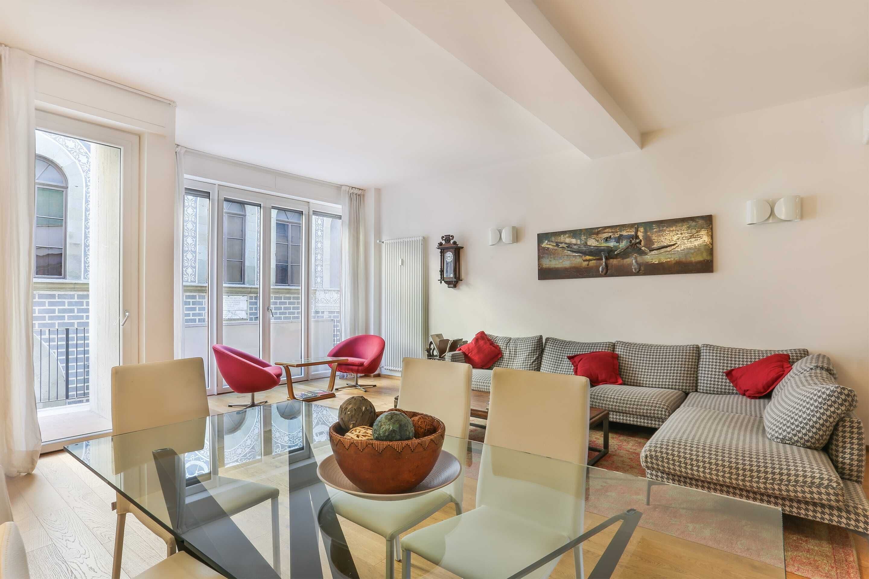 A modern living room with a glass dining table, red chairs, a patterned sofa, and a large window with a view of the city.