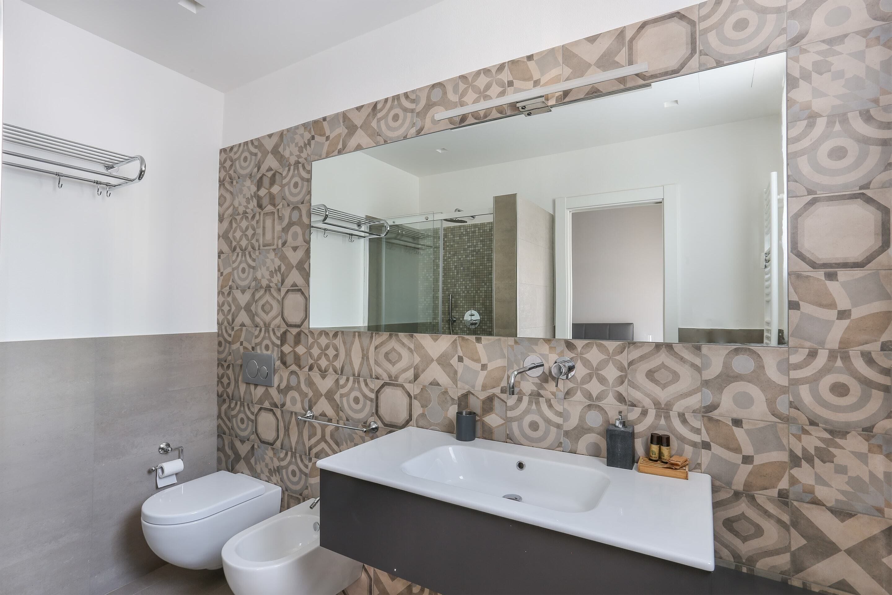A modern bathroom with a large mirror, a white sink, a bidet, and patterned tiles.