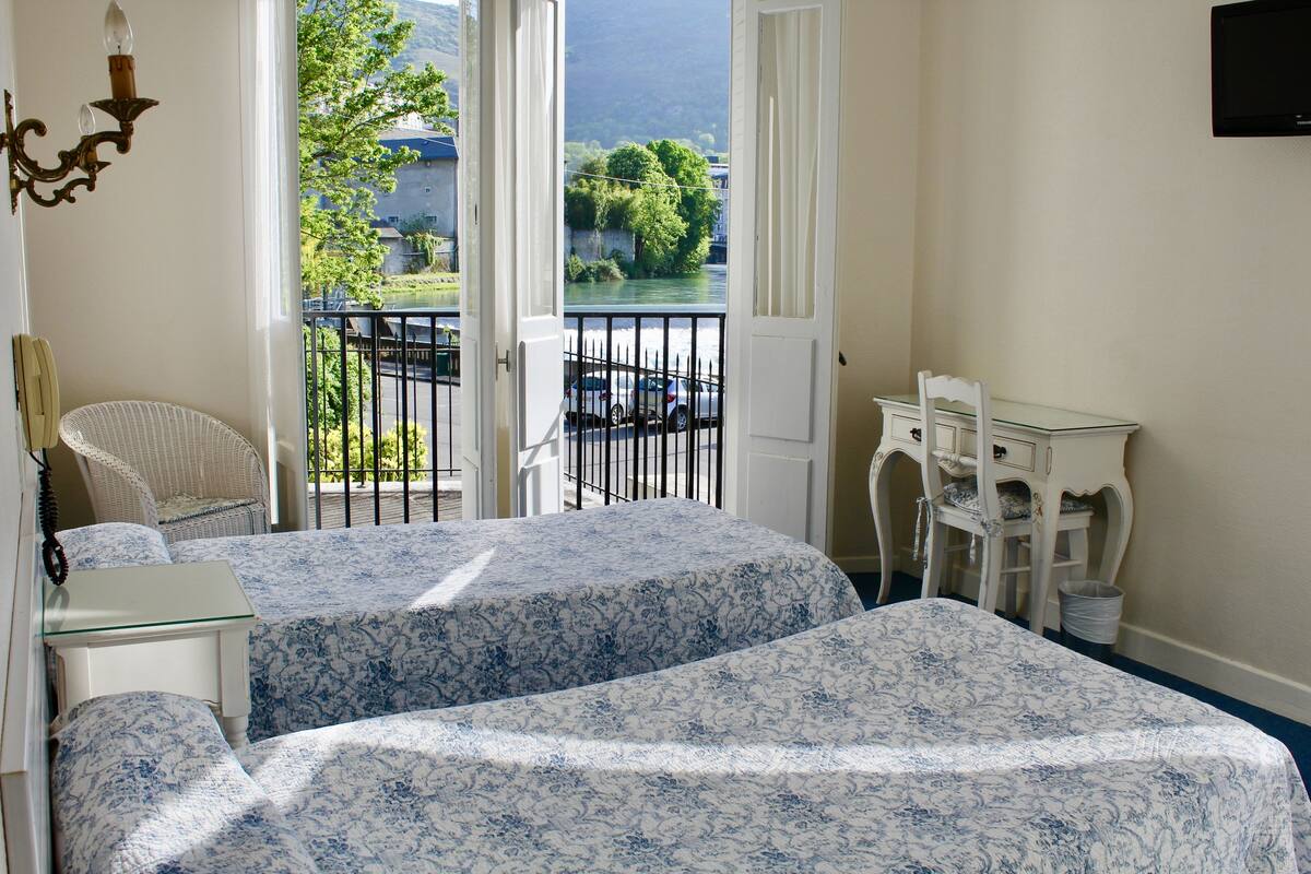 A bedroom with two beds, a chair, a desk, and a view of a balcony with a railing and a car parked outside.