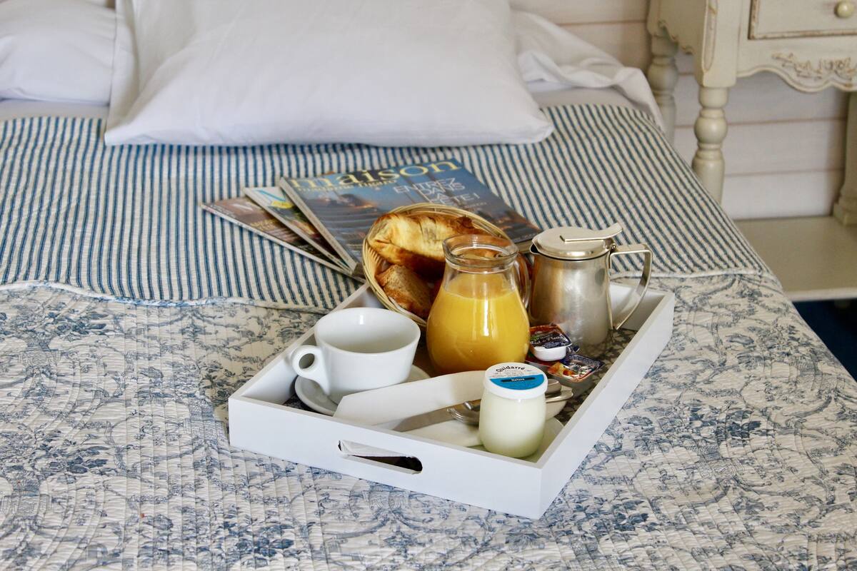 A breakfast tray with a cup, a glass of orange juice, a bottle of milk, and a jar of jam on a bed with a striped bedspread and a magazine.