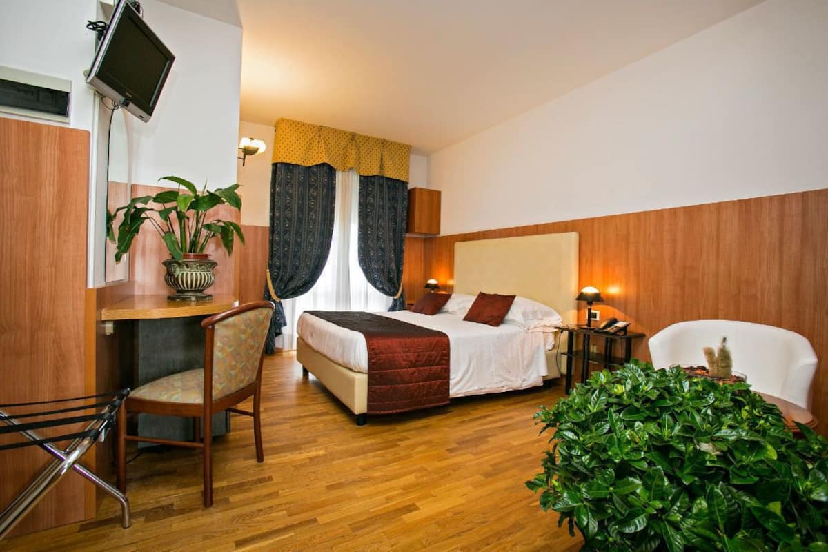 Ein Hotelzimmer mit einem Bett, einem Schreibtisch mit Stuhl, einem Fernseher, einer Topfpflanze und einer großen Zimmerpflanze.