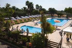 Un área de piscina con sillas de descanso y sombrillas, rodeada de palmeras y follaje.