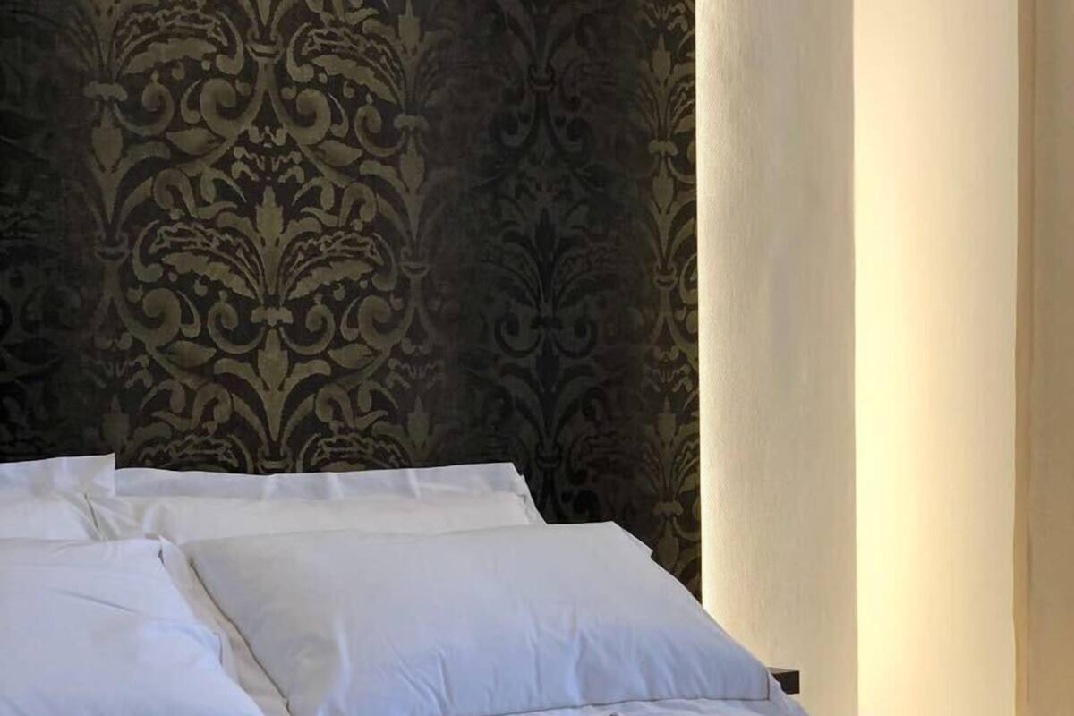 A neatly made bed with white linens and a black accent, set against a patterned wallpaper.