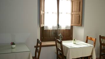 Un comedor con una mesa y sillas, una ventana con contraventanas de madera y una planta en maceta sobre una mesita.
