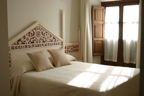 A bedroom with a bed, two pillows, a wooden headboard with intricate patterns, a window with curtains, and a wooden door. - Santa María de Úbeda (Úbeda)